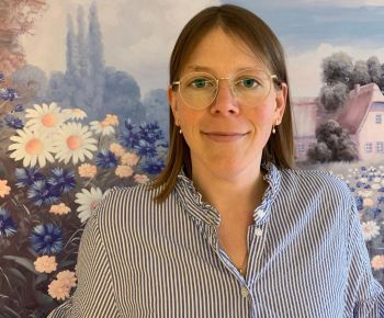Eine Frau mit Brille lächelt vor einem malerischen Blumenhintergrund und einem idyllischen Haus in der Landschaft.