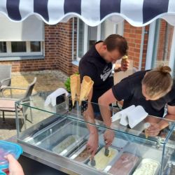 Unter einem gestreiften Vordach servieren zwei Männer Eis aus einer Kühltheke an wartende Kunden.