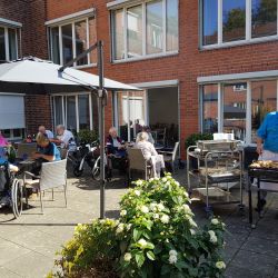 Ein Koch grillt Fleisch im sonnigen Garten eines Seniorenheims, während ältere Menschen gemütlich im Freien plaudern.