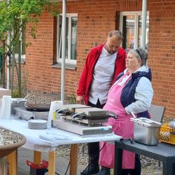 Zwei Personen unterhalten sich neben einem Tisch mit Küchengeräten vor einem Backsteinhaus im Freien.