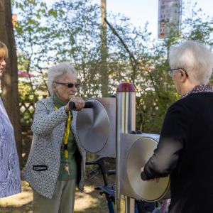 Drei Frauen interagieren mit einem Spielgerät im Freien, umgeben von Bäumen und einem sonnigen Hintergrund.