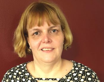 Eine Frau mit kurzem, blondem Haar lächelt in einem gepunkteten Oberteil vor einer leuchtend roten Wand.