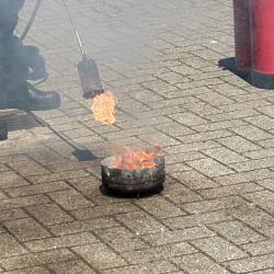 Ein roter Feuerlöscher steht neben einem brennenden Behälter, während eine Person mit einer Flamme auf dem Boden arbeitet.