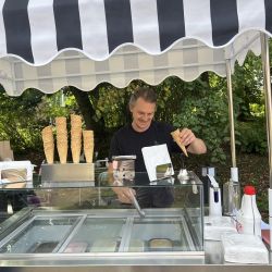 Ein Mann serviert Eis aus einem mobilen Eiswagen mit schwarz-weiß gestreiftem Dach in einer grünen Umgebung.