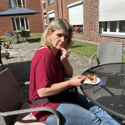 Eine Frau in rotem Oberteil genießt bei Sonnenschein ein Stück Kuchen an einem Metalltisch im Freien.
