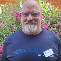 Ein lächelnder Mann mit Brille steht vor blühenden Pflanzen und trägt ein T-Shirt mit der Aufschrift "PUSH THE LINE".