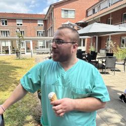 Ein Mann in grüner Arbeitskleidung genießt ein Eis im sonnigen Innenhof eines roten Backsteingebäudes.