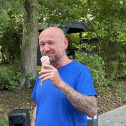 Ein Mann mit Tätowierungen genießt an einem sonnigen Tag im Grünen ein Eis, umgeben von üppigen Bäumen und Pflanzen.