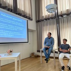 Drei Männer sitzen in einem Raum und verfolgen aufmerksam eine Präsentation über Maßnahmen gegen Gewalt auf einer Leinwand.