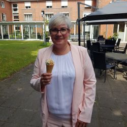 Eine Frau in einem hellrosa Anzug genießt ein Eis auf einem gepflasterten Hof vor einem Backsteingebäude.