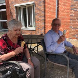 Ein älteres Paar sitzt entspannt vor einem Backsteinhaus und genießt Eis in der warmen Sonne.