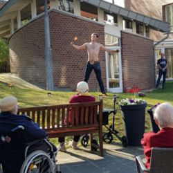 Ein Mann jongliert vor einem modernen Backsteingebäude und unterhält eine Gruppe von Senioren im Freien.