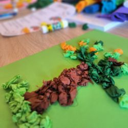 Bunte Papierstücke bilden einen Baum auf grünem Papier, umgeben von Bastelmaterialien auf einem Tisch.