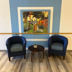 Zwei blaue Sessel flankieren einen runden Tisch vor einem farbenfrohen Gemälde an der blau umrahmten Wand.
