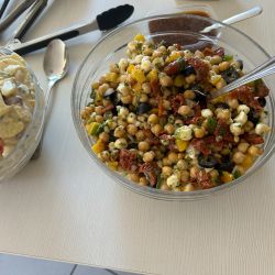 Ein bunter Kichererbsensalat mit getrockneten Tomaten und Oliven steht neben einem cremigen Nudelsalat auf einem Tisch.