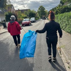 Zwei Personen gehen die Straße entlang und sammeln mit blauen Tüten und Greifzangen Müll auf.