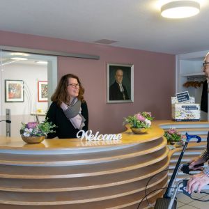 Ein älterer Mann mit Gehhilfe spricht lächelnd mit einer Empfangsdame in einem einladenden Foyer. | © Name