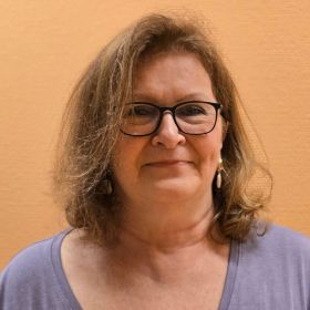 Eine Frau mit Brille und lockigem Haar lächelt vor einer orangefarbenen Wand. | © Name
