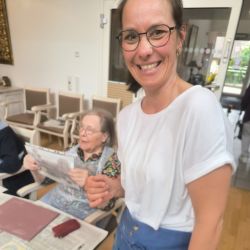 Eine lächelnde Frau hält die Hand einer älteren Dame, während sie in einem hellen Raum mit Kunstmaterialien sind.