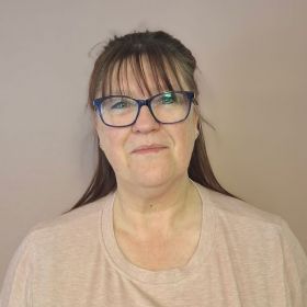 Eine Frau mit Brille und langen Haaren schaut freundlich in die Kamera vor einem neutralen Hintergrund.