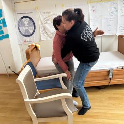 Zwei Frauen üben Kinästhetik neben einem Krankenbett, umgeben von Lehrmaterialien und Diagrammen an der Wand.