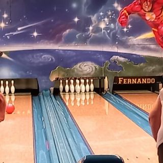 Zwei Personen bowlen vor einer bunten Wand mit Superhelden-Motiven und rollen die Kugel gezielt auf die Pins.