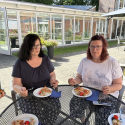 Zwei Frauen sitzen an einem runden Tisch im Freien und genießen Pizza unter einem Sonnenschirm.