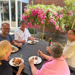 Fünf Personen genießen ein Mittagessen im Freien neben leuchtend rosa Blumen an einem sonnigen Tag.