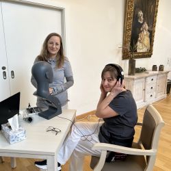 Zwei Frauen arbeiten an einem Tisch im Büro, eine trägt Kopfhörer, während eine dekorative Büste daneben steht.