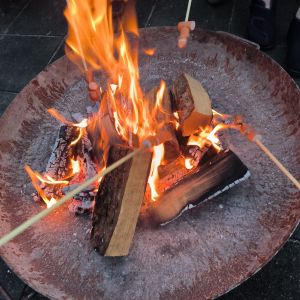 Mehrere Stockbrote werden über einem lodernden Lagerfeuer in einer Feuerstelle geröstet, während Flammen aufsteigen.