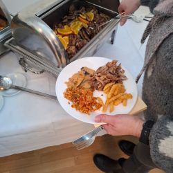 Eine Person serviert sich an einem Buffet einen Teller mit Fleisch, Pommes und Reis.
