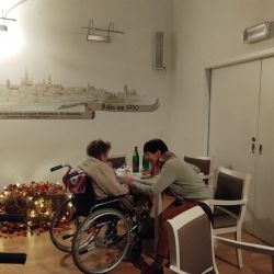 Eine Frau unterhält sich liebevoll mit einem Rollstuhlfahrer in einem gemütlichen Raum mit herbstlicher Dekoration.