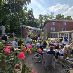 Im Garten eines Seniorenheims genießen Menschen bei sonnigem Wetter geselliges Beisammensein, umgeben von blühenden Rosen.