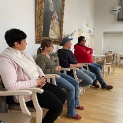 Vier Personen sitzen aufmerksam in einem Raum mit Holzboden, während ein großes Gemälde an der Wand hängt.
