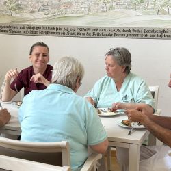 Mehrere Personen in Arbeitskleidung genießen gemeinsam ihre Pause beim Essen an einem Tisch in einem hellen Raum.