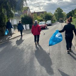Eine Gruppe von Menschen sammelt Müll mit blauen Säcken in einer Wohnstraße unter einem bewölkten Himmel.