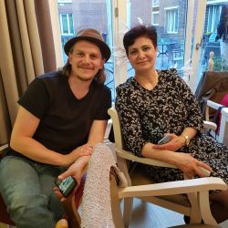 Ein Mann und eine Frau sitzen lächelnd in einem gemütlichen Raum auf Stühlen, umgeben von Luftballons und halten Smartphones.