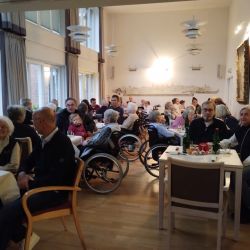 In einem gemütlichen Raum unterhalten sich viele Menschen, darunter ältere Personen, lebhaft an Tischen.