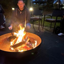 Ein Mann kniet am Abend neben einem Lagerfeuer, umgeben von Gartenstühlen und Bäumen im Freien.