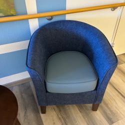 Ein blauer Sessel mit grauem Sitzkissen steht auf einem Holzfußboden neben einem runden Tisch vor einer blauen Wand.