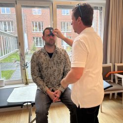 In einem hellen Raum misst ein Mann mit einem blauen Werkzeug die Stirn eines sitzenden Mannes.