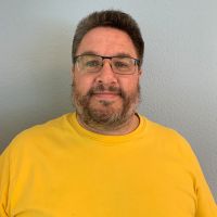 Eine Person mit Brille und Bart trägt ein gelbes T-Shirt vor einem hellen Hintergrund.