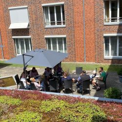 Eine Gruppe von Menschen sitzt entspannt unter Sonnenschirmen auf einer Terrasse vor einem Backsteingebäude und genießt den Tag.