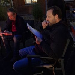 Zwei Männer sitzen draußen; einer liest aufmerksam ein Dokument, während er einen Snack genießt.