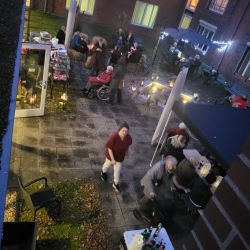 Eine Gruppe von Menschen genießt einen gemütlichen Abend auf einer beleuchteten Terrasse mit Tischen, Lichtern und Getränken.