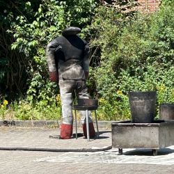 Ein verlassener Schweißanzug steht neben einem Metallbehälter, umgeben von grünen Büschen und einem Ziegelgebäude im Freien.