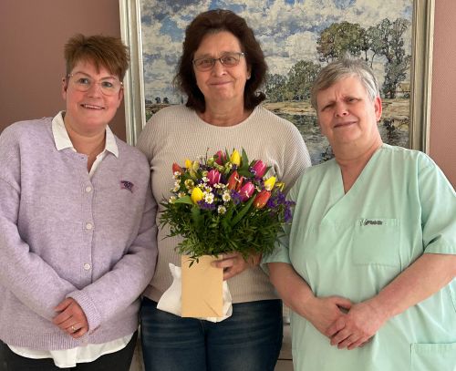 Drei Frauen lächeln stolz, während eine von ihnen einen bunten Blumenstrauß in der Hand hält.
