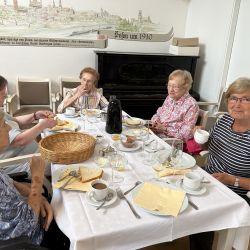 Eine Gruppe älterer Frauen genießt Kaffee und Kuchen an einem Tisch mit einem historischen Wandbild im Hintergrund.