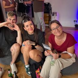 Drei Personen sitzen entspannt auf dem Boden, lächeln in die Kamera und genießen Getränke in einer lockeren Atmosphäre.