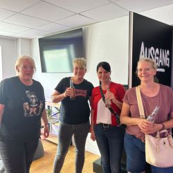 Vier Frauen stehen lächelnd auf einem Holzboden, halten Getränke und posieren neben einem Schild mit der Aufschrift "Ausgang".